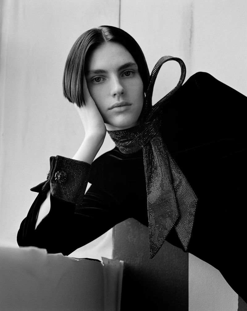 Monochrome portrait of a fair-skinned adult with short dark hair wearing a black velvet dress and sequined necktie against a neutral backdrop.