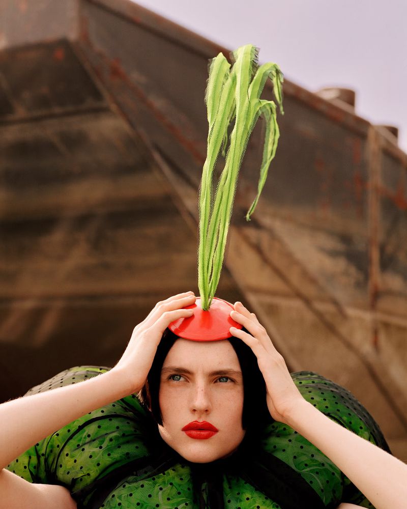 Artistic fashion portrait: Woman with dark hair, red lipstick, wearing green dress and red hat with green sprouts, against blurred metal background.