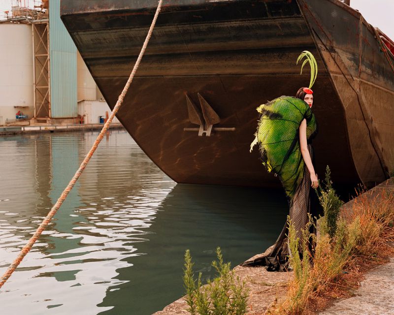 Fashion shot of a woman in an avant-garde green costume posing by the water near a weathered ship, creating a striking contrast of textures.