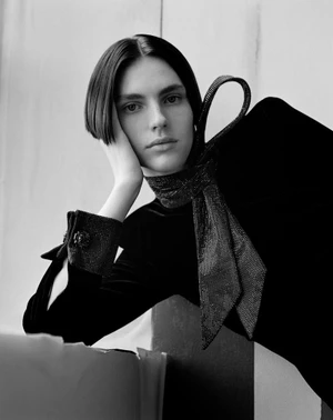 Monochrome portrait of a model with short dark hair leaning their head on their hand, wearing a dark velvet top and sparkling bow tie.