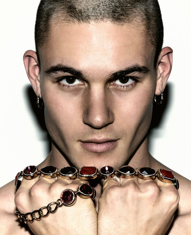 Close-up of a young adult man with a buzz cut, wearing statement jewelry: a bracelet and a ring with large, dark gemstones. Indoor shot.