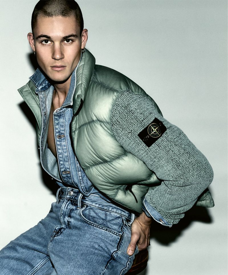 Male fashion model poses in a denim shirt and jeans, accented by a light green puffer jacket, against a neutral backdrop.