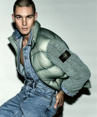 Male fashion model poses in a denim shirt and jeans, accented by a light green puffer jacket, against a neutral backdrop.