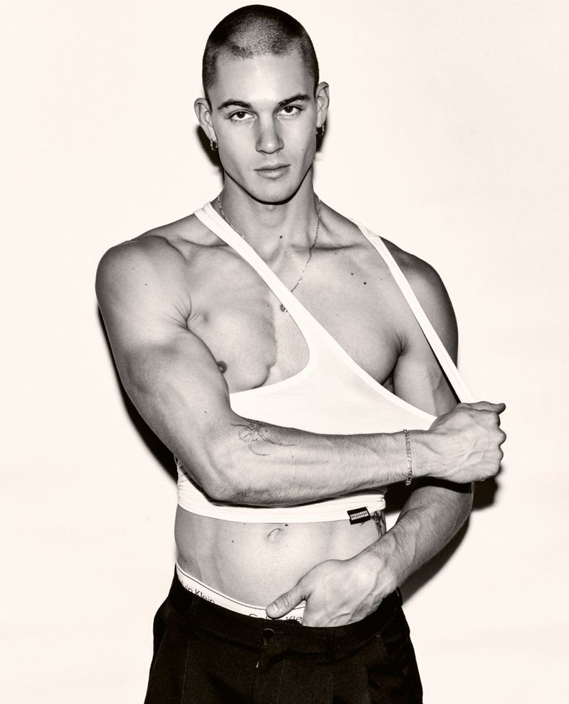 Masculine male model poses indoors in a white tank top and dark pants against a plain background, creating a high-contrast portrait.