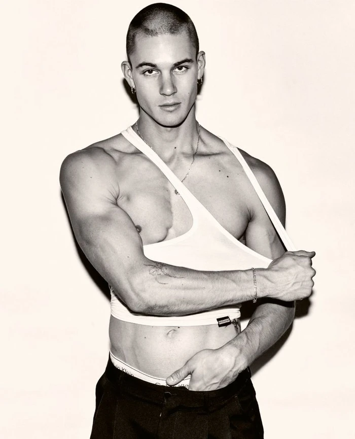 Model Samuel Watson wearing a white tank top pulled off one shoulder and black trousers against a pale studio backdrop.