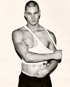 Model Samuel Watson wearing a white tank top pulled off one shoulder and black trousers against a pale studio backdrop.