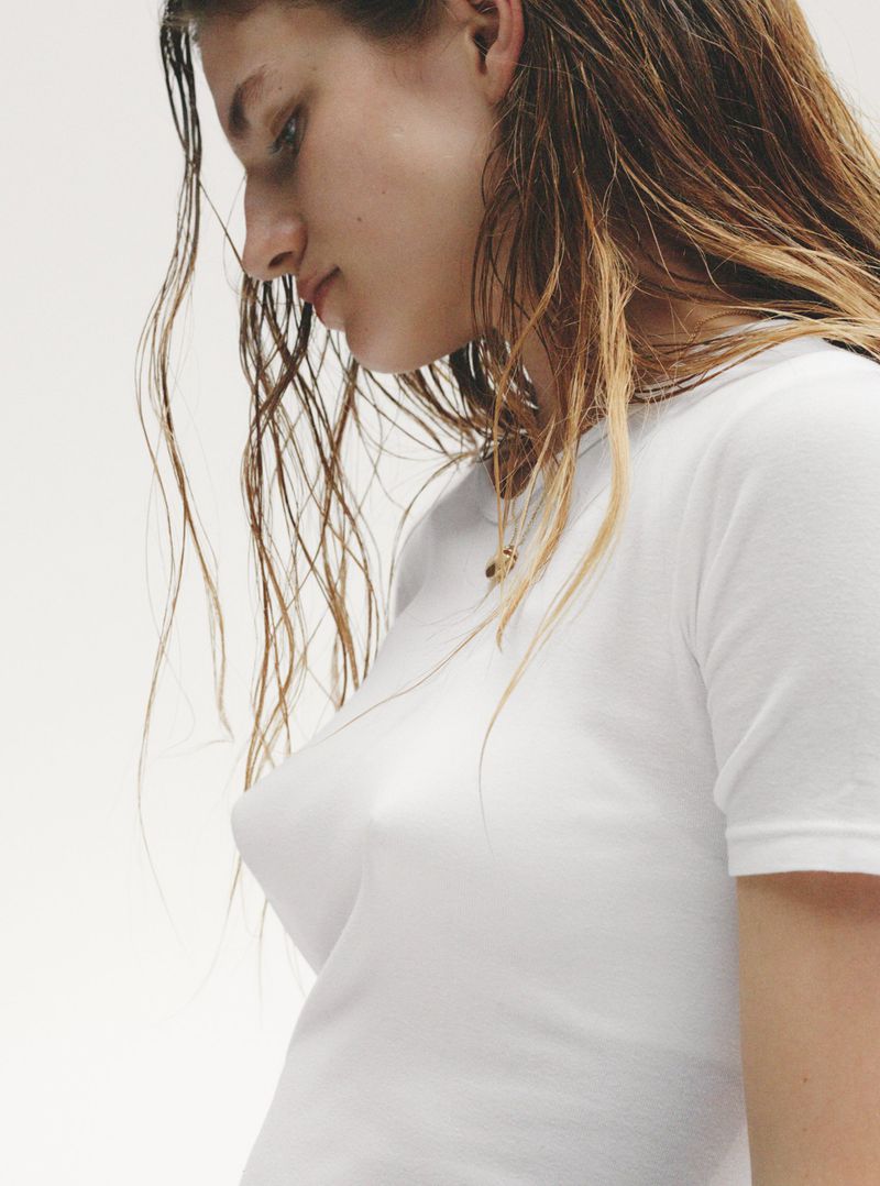 Close-up of a young woman with wet brown hair wearing a white t-shirt against a plain white background, looking down with a neutral expression.