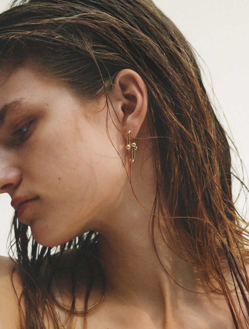 Close-up shot of a light-skinned woman with wet brown hair wearing gold dangling earrings against a neutral background.