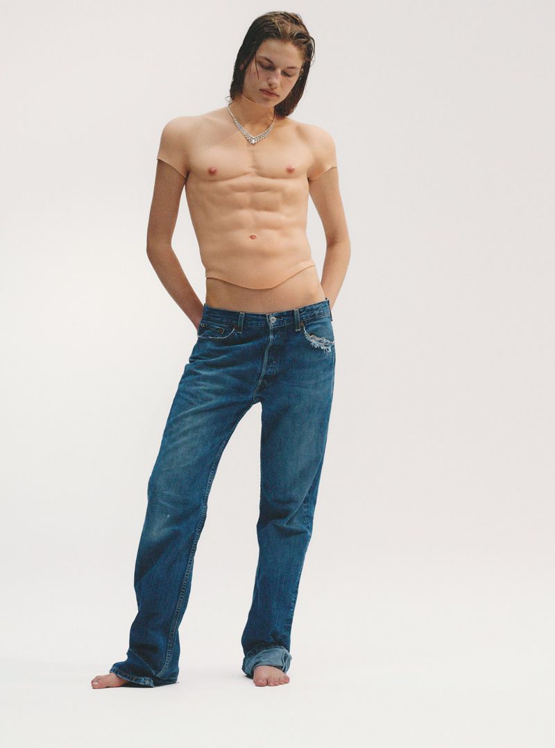 A slender, fair-skinned person stands in a photo studio wearing jeans and a prosthetic torso with imitation abs and chest, shot in a studio.