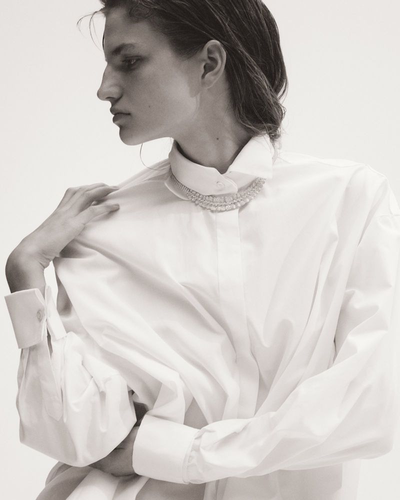 Monochrome portrait of an adult woman wearing a white button-down shirt and a beaded necklace, hand on her shoulder, against a white backdrop.