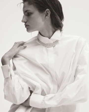 Monochrome portrait of an adult woman wearing a white button-down shirt and a beaded necklace, hand on her shoulder, against a white backdrop.