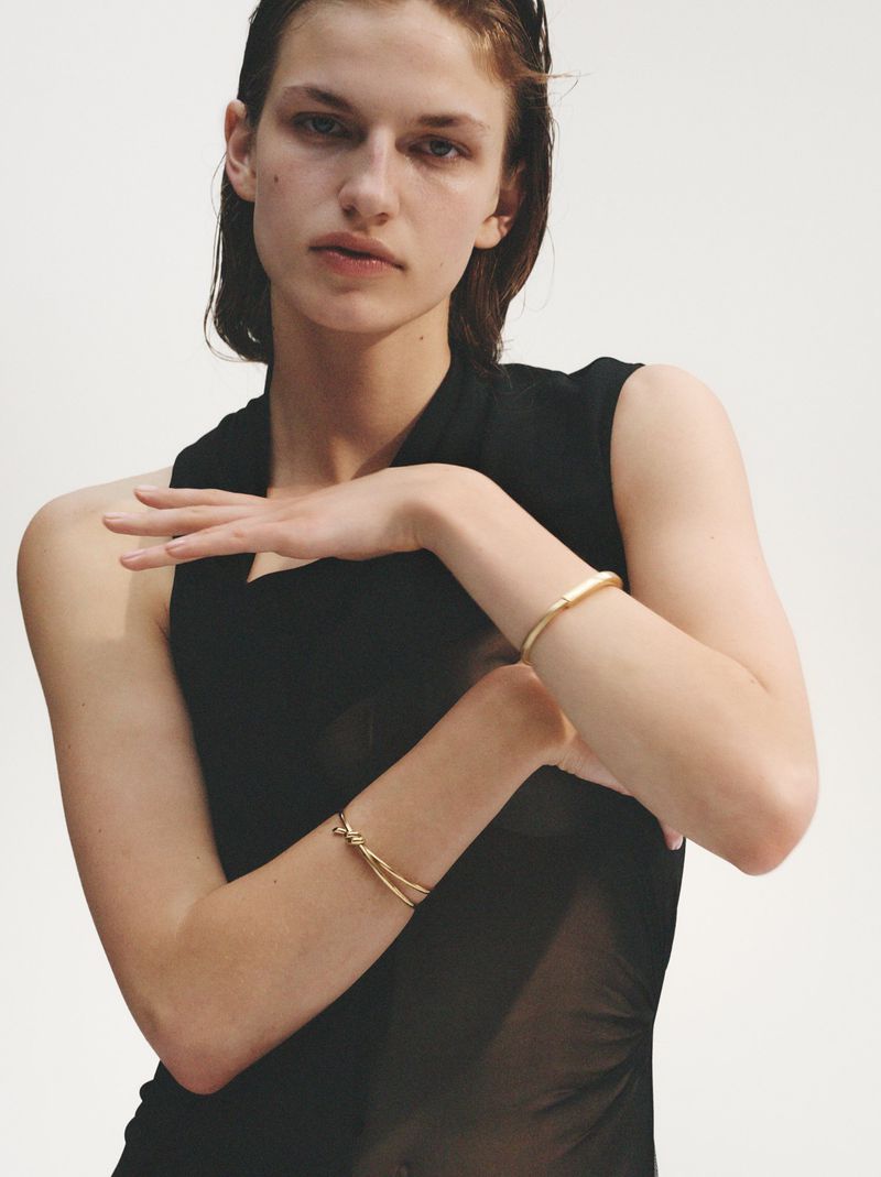 Close-up of a young woman in a black dress, posing with arms raised to display golden bracelets on both wrists against a bright backdrop.