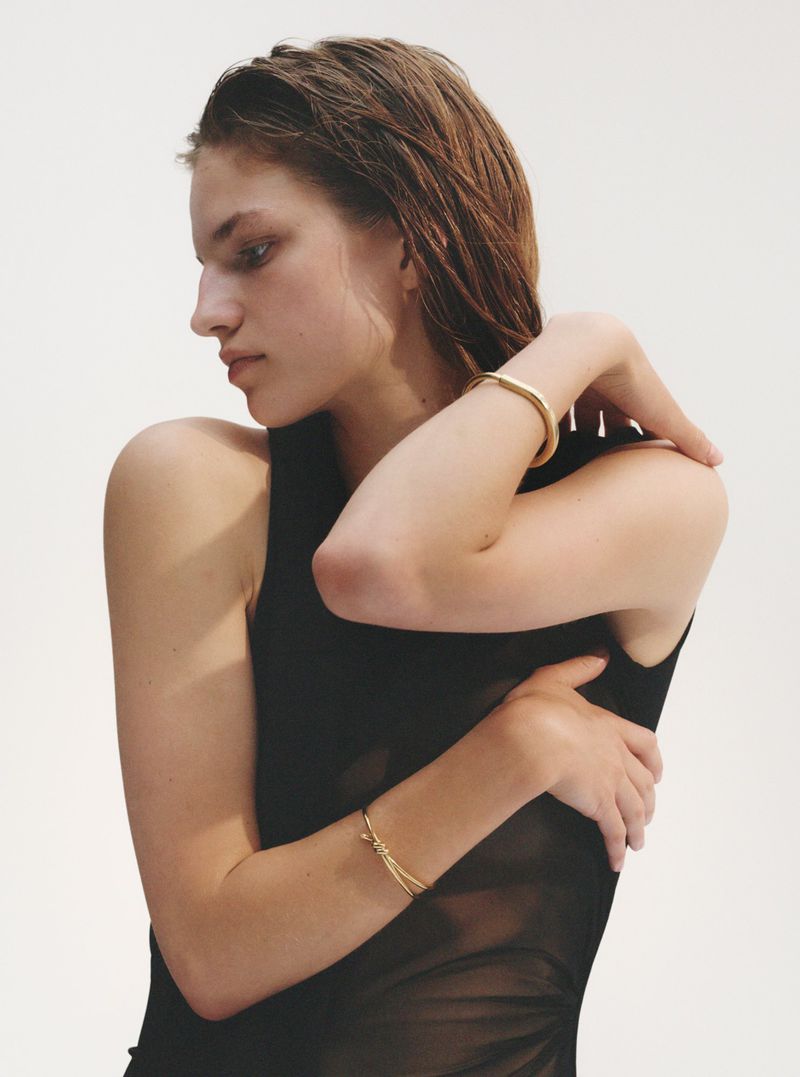 Model poses in a sleeveless black top with gold bracelets, shot in a photo studio against a white backdrop with soft lighting.