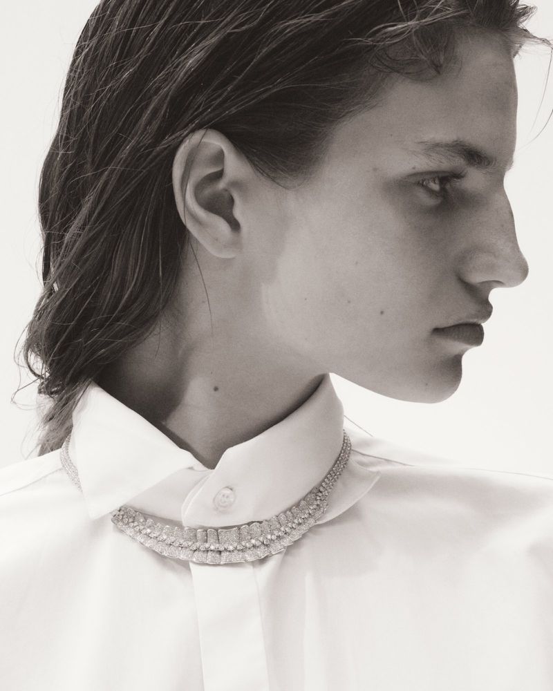Monochrome close-up studio portrait of an adult female model wearing a white collared shirt and necklace against a bright white background.