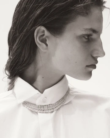 Monochrome close-up studio portrait of an adult female model wearing a white collared shirt and necklace against a bright white background.