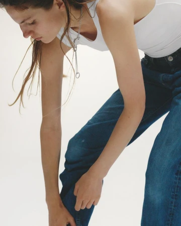 Young woman in white tank top and jeans poses against white backdrop. Fashion shot with natural light and minimalist styling.