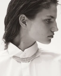 Model in profile wearing a white collared shirt with a layered crystal necklace around the collar against a white background.