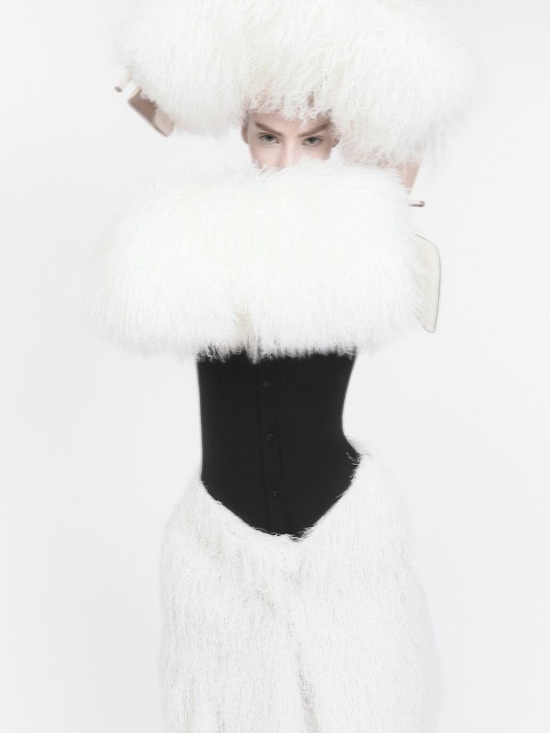 Fashionable person with a large white fluffy hat and matching outfit poses in a studio setting with pale, diffused lighting.