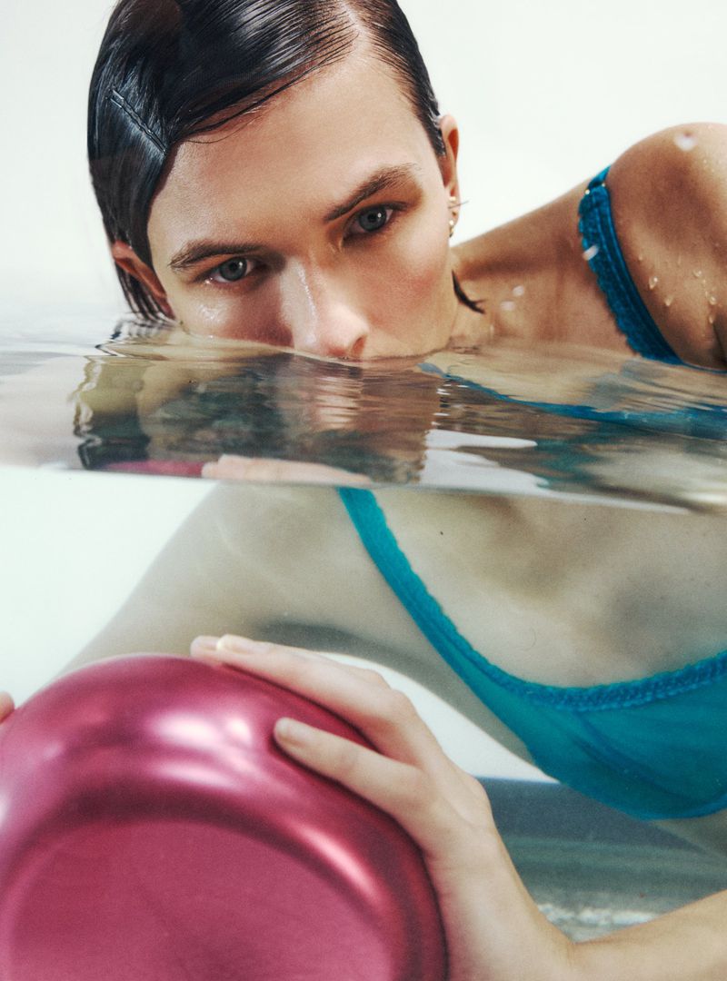 Person submerged in water, chest-up, holding a pink object; wears a turquoise bra and has wet dark hair slicked back. Studio setting.