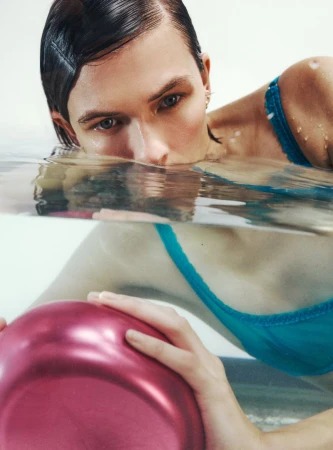 Person submerged in water, chest-up, holding a pink object; wears a turquoise bra and has wet dark hair slicked back. Studio setting.