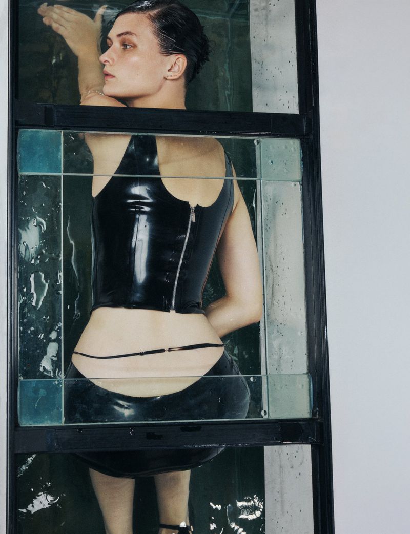 Woman in a bathtub with a black latex top and bottom, shot through an ornate glass partition, creating a stylized and fashionable portrait.