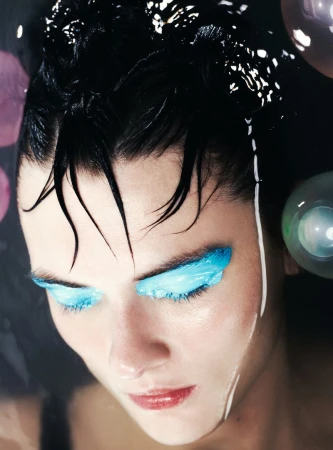 Close-up portrait of a woman in water with bright blue eyeshadow, showcasing beauty, makeup, and dark hair against a dark background.