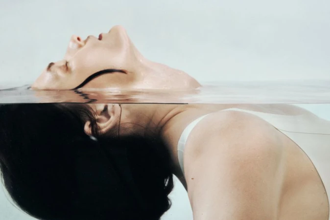 Adult woman in water with head tilted back, partially submerged and reflecting, lit with soft light against a pale background.