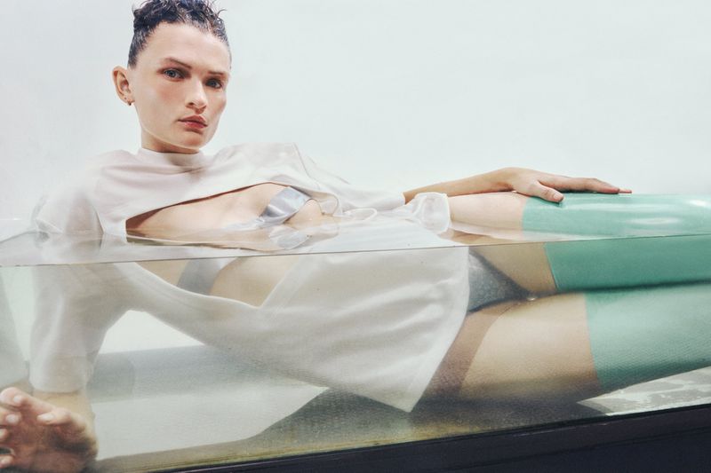 Adult posing in water tank wearing a white crop top, silver tape, and dark underwear. Horizontal shot with studio lighting.