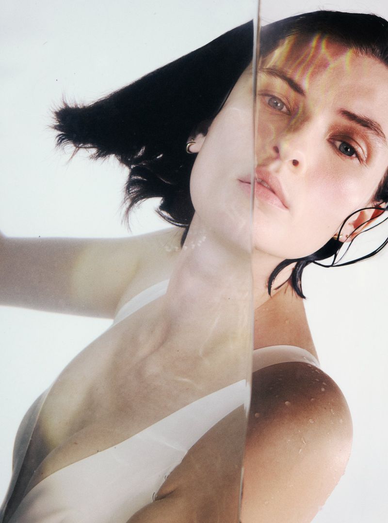 Woman with dark hair and wet skin poses behind rippled glass in an eye-level close-up shot in a photo studio with bright lighting.