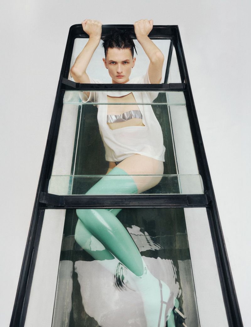 Androgynous model poses inside a glass structure wearing a white t-shirt, mint thigh-high stockings, and heeled boots against a white backdrop.