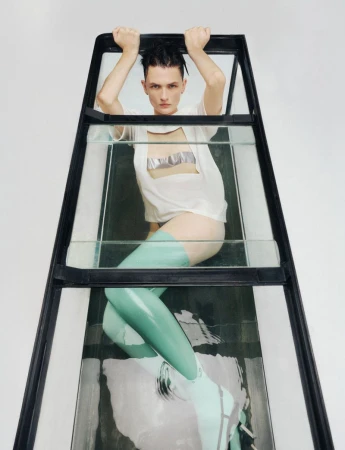 Androgynous model poses inside a glass structure wearing a white t-shirt, mint thigh-high stockings, and heeled boots against a white backdrop.