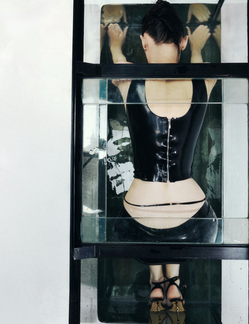 Woman in a black corset submerged in water behind a glass frame, viewed from the rear, with high-heeled shoes at the bottom.