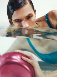 Model Lara Mullen partially submerged in water, looking at the camera while holding a metallic pink bowl.
