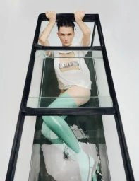 Model Lara Mullen lying inside a narrow glass enclosure wearing a white cutout shirt and mint green latex stockings on white seamless.
