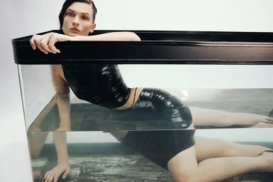 Model Lara Mullen wearing black latex apparel lying inside a glass water tank against a white background for HardCOPY Magazine