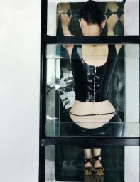 Model Lara Mullen viewed from behind in a black zip-back latex top and heels inside a glass water tank setup.
