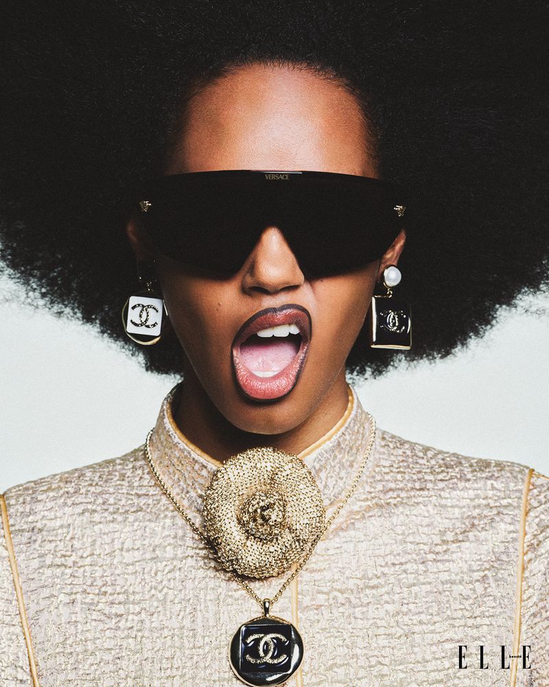 Fashion editorial shot of a Black woman wearing designer sunglasses, jewelry, and a metallic gold jacket, with an open mouth.