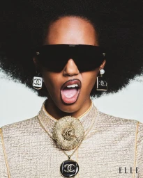 Model Sabryna Oliveira posing with an open mouth wearing black shield sunglasses, Chanel earrings, and a gold top against white.