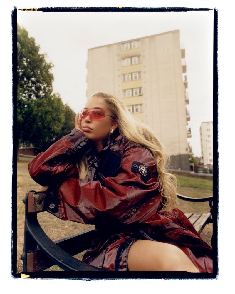 Woman with blond hair sitting on a park bench, wearing a red jacket and red sunglasses with a tall building in the background.