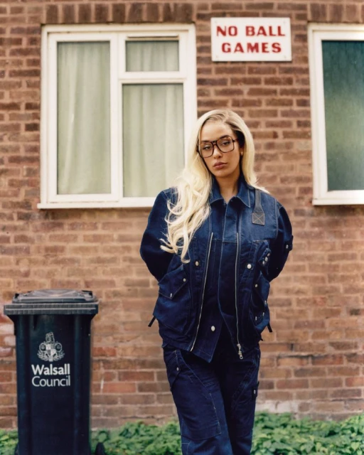 A woman with long blonde hair poses in front of a brick building with a 'No Ball Games' sign and a trash can.