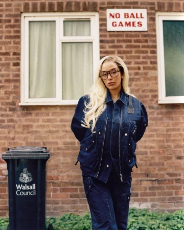 A woman with long blonde hair poses in front of a brick building with a 'No Ball Games' sign and a trash can.
