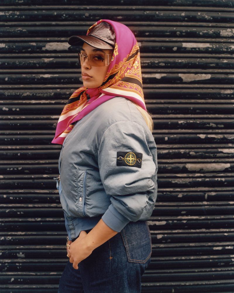 Woman with translucent glasses, headscarf, and baseball cap posing in front of a corrugated metal wall in a medium shot.