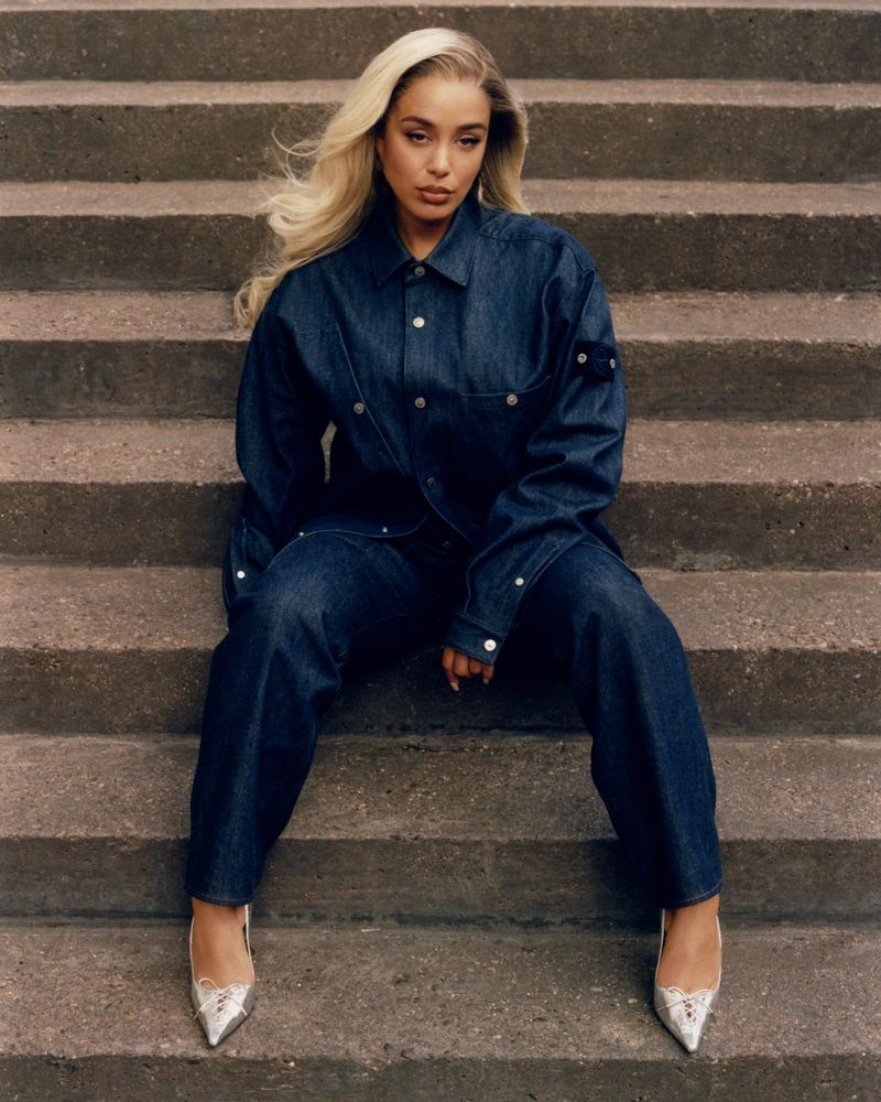 A woman in a denim outfit poses on concrete steps. She is wearing snakeskin heels and has long blonde hair styled with a middle part.