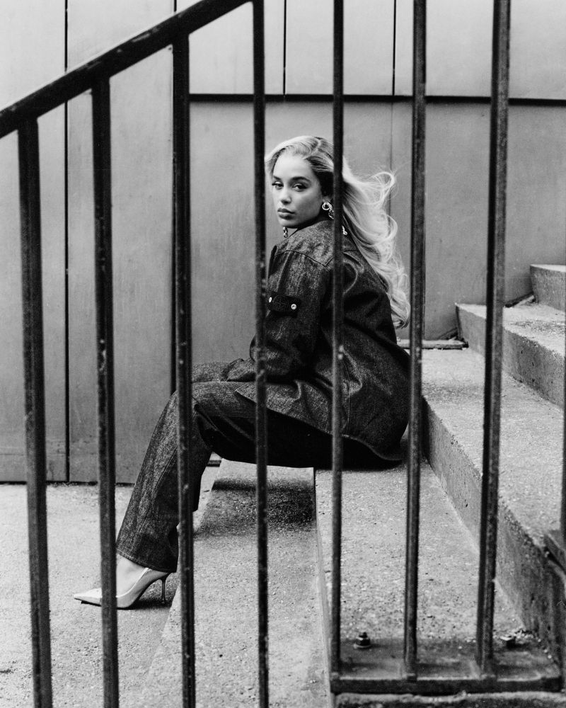 Monochrome fashion portrait of a blonde woman in a denim suit sitting on concrete steps behind a metal railing, looking at the camera.