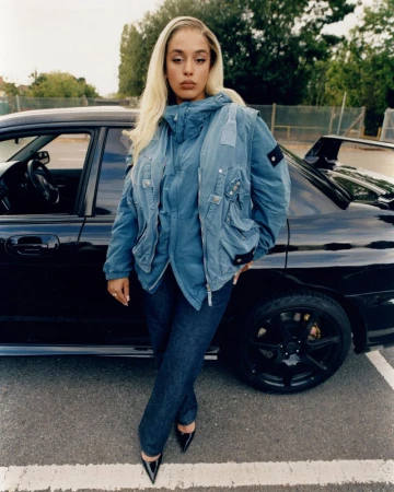 A woman with long blonde hair poses next to a black sports car in an outdoor setting, wearing a blue jacket, jeans, and black high heels.