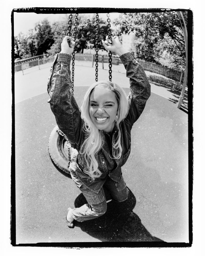 Monochrome shot of a woman in a denim outfit smiling with one eye closed while on a tire swing in an outdoor park.