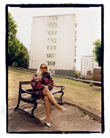 A woman with long blond hair and sunglasses sits on a park bench in front of a tall apartment building in an urban outdoor scene.