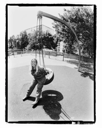 A woman with long blonde hair swings in a tire swing in a black-and-white photo taken at eye-level on a sunny day in an outdoor park.