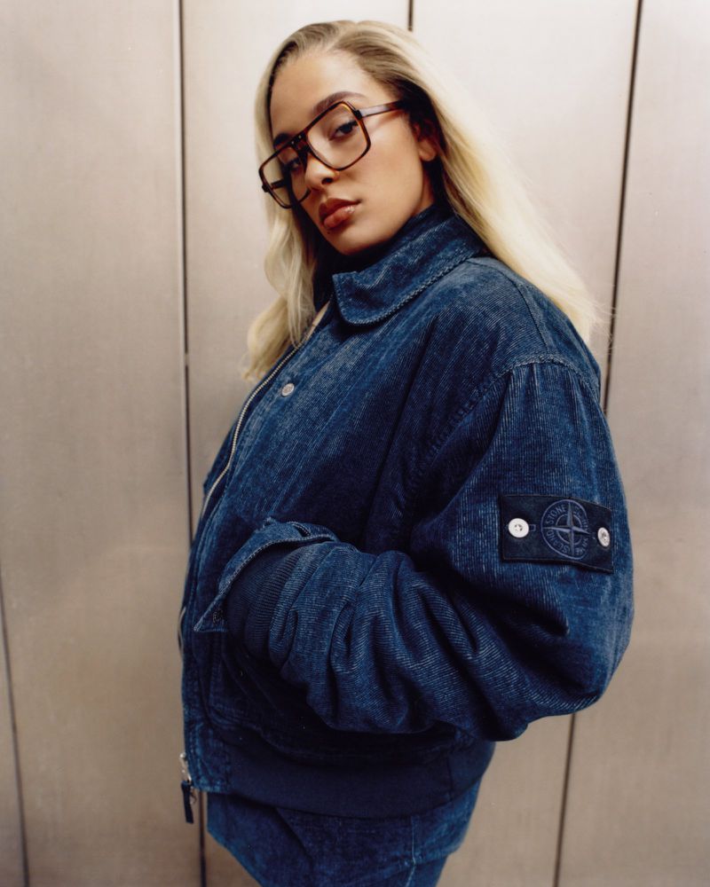 Portrait of a young adult woman with blonde hair wearing a denim jacket and glasses, posing indoors against a gray wall.