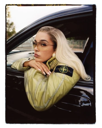 Close-up of a woman with platinum blonde hair wearing sunglasses and a lime green jacket sitting inside of a black car.